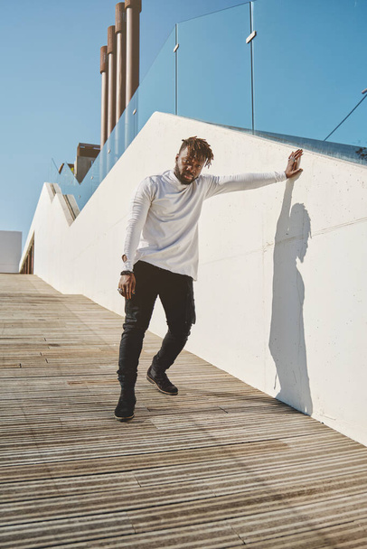 Full body of young African American male in trendy casual clothes standing on wooden ramp and leaning on concrete wall against blue sky while looking at camera - Photo, Image