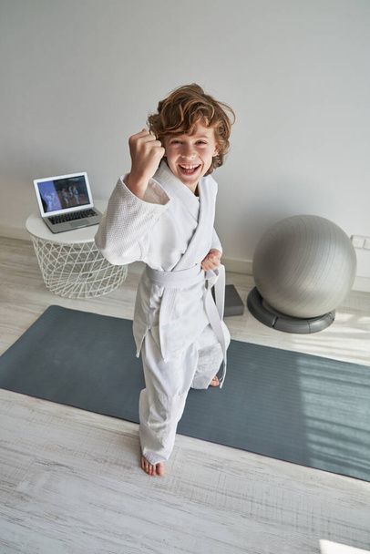 Full body of positive curly haired preteen kid in white kimono standing on yoga mat with clenched fists while practicing karate with online lesson on laptop - Photo, Image