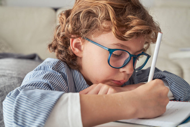 Günlük giysiler ve gözlükler içinde konsantre olmuş çocuk aşağı bakarken ve ödevlerini yaparken not alırken masada oturuyor. - Fotoğraf, Görsel