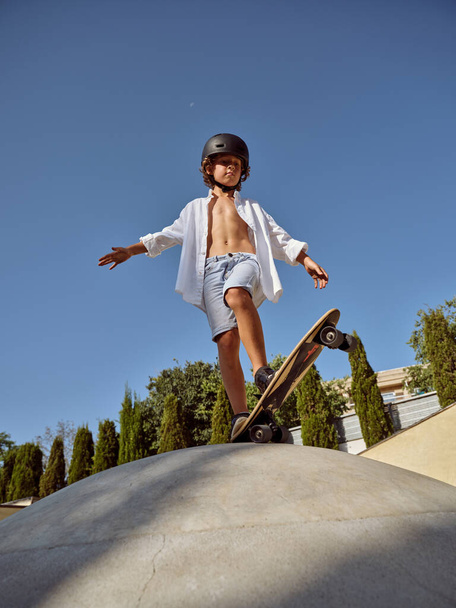 Cuerpo completo de niño en traje casual y casco mirando a la cámara mientras está de pie en el monopatín contra el cielo azul sin nubes - Foto, Imagen