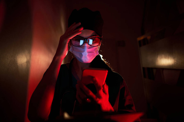 Обеспокоенная женщина-врач в медицинской форме и защитной маске сидит на лестнице и трогает лоб во время просмотра смартфона в темной комнате при красном освещении - Фото, изображение