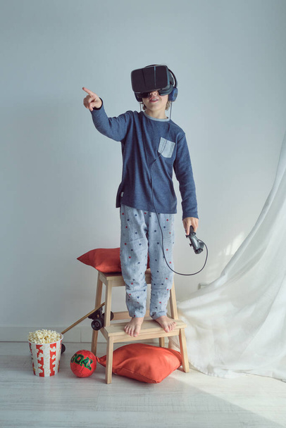 Full body of barefoot preteen boy in pajamas standing on stool stepladder in virtual reality headset and headphones and with joystick and pointing away with forefinger - Photo, Image