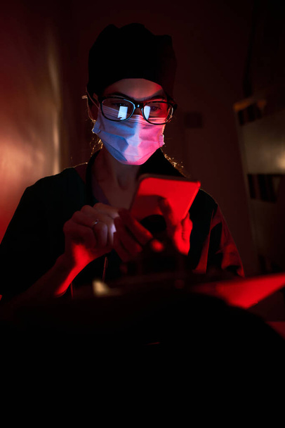 Besorgte erwachsene Ärztin in Arztuniform und Schutzmaske sitzt auf der Treppe und berührt die Stirn, während sie im dunklen Raum unter rotem Licht mit dem Smartphone surft - Foto, Bild