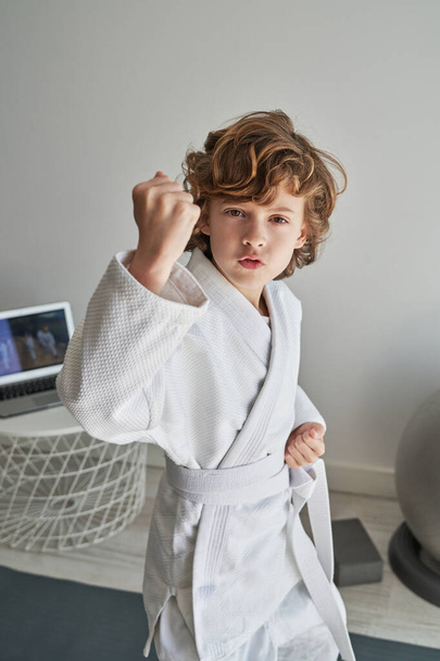Beyaz kimonolu, kemerli, dudak büken, judo çalışırken kameraya bakan ve evde yumruk sıkan ciddi bir çocuk. - Fotoğraf, Görsel
