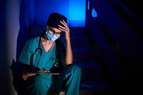 Nachdenkliche Sanitäterin in Schutzmaske und Uniform sitzt auf der Treppe mit Klemmbrett, während sie Dokumente liest und den Kopf berührt - Foto, Bild