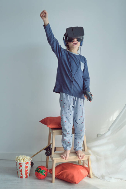 Full body of anonymous barefoot kid in pajama raising arm with open mouth while playing virtual reality game wearing headset and standing on ladder against white wall - Photo, Image