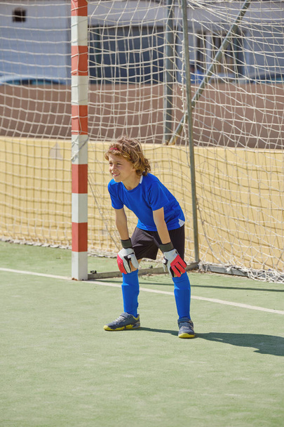 Πλήρες σώμα του προ-έφηβος τερματοφύλακας σε activewear κλίνει στα γόνατα και ατενίζοντας προς τα εμπρός, ενώ παίζει ποδόσφαιρο κατά τη διάρκεια της κατάρτισης σε γήπεδα - Φωτογραφία, εικόνα