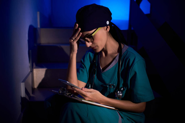 Médico femenino confuso en uniforme médico con estetoscopio sentado en la escalera con portapapeles y el teléfono móvil de navegación en la oscuridad bajo iluminación azul - Foto, imagen