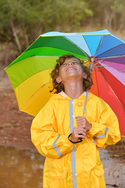 Niño alegre con paraguas de arco iris vistiendo impermeable amarillo de pie con los ojos cerrados en charco en el bosque - Foto, imagen