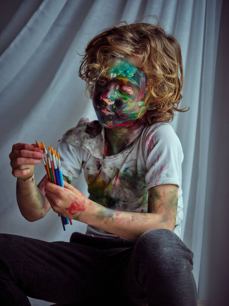 Хлопчик в повсякденному вбранні, покритий барвистими фарбами, вивчає набір пензлів в руці, сидячи в світлій кімнаті - Фото, зображення