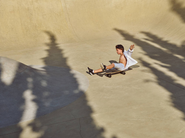Вид сбоку на маленького мальчика в повседневной одежде, сидящего на скейтборде верхом в скейт-парке с поднятыми руками, балансируя и практикуясь в одиночку - Фото, изображение