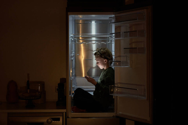 自宅でダークキッチンでタブレットを交差し,使用して足で冷蔵庫に座っているカジュアルな服の深刻な男の子の側面 - 写真・画像
