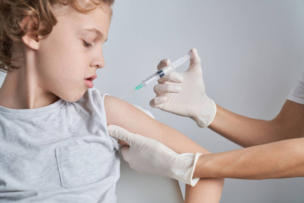 Crop unrecognizable doctor in latex gloves injecting vaccine into shoulder of boy on white background during epidemic in light room - Photo, Image