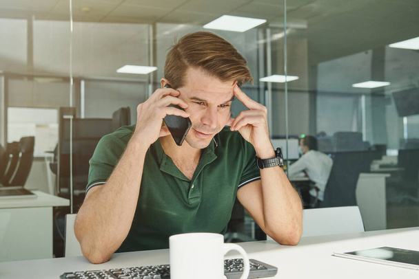 Pracownik biurowy w zielonej koszuli rozmawiający przez telefon komórkowy siedząc przy stole w nowoczesnym miejscu pracy - Zdjęcie, obraz