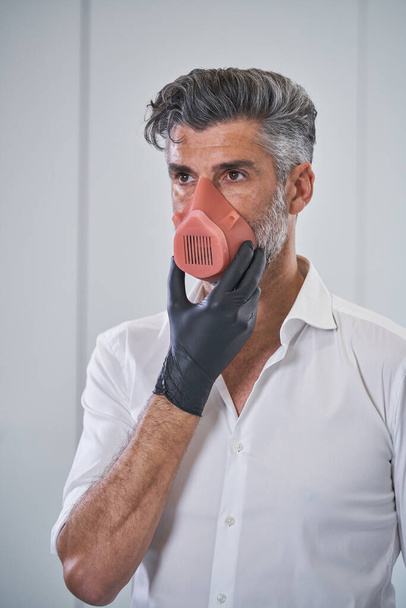 Serious bearded male wearing respirator and medical gloves touching chin and preventing infection spread while standing on white background - Photo, Image