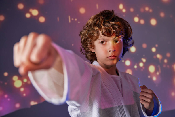 Serious sportive boy with curly hair in white kimono doing punch during martial judo training in studio with bright shimmering lights - Photo, Image
