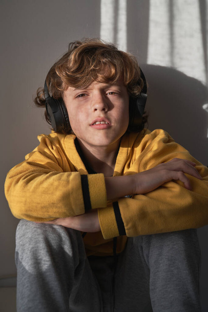 Seriöser Junge in lässiger Kleidung, der in die Kamera schaut, während er Musik in drahtlosen Kopfhörern hört und allein zu Hause sitzt - Foto, Bild