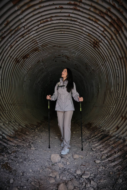 Full body of Hispanic female with trekking poles looking up while observing shabby rough wall in long old tunnel during trip - Photo, Image