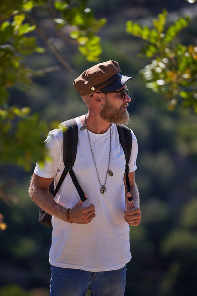 Bearded male tourist in sunglasses and cap with backpack standing near green trees against mountain slope and looking away - Photo, Image