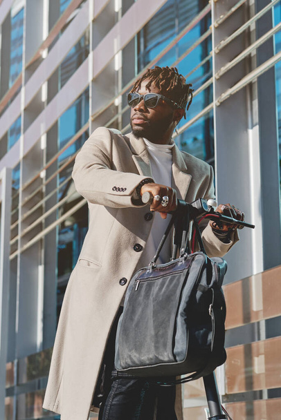 Low angle of African American bearded male in casual outfit and sunglasses standing with handbag in sunlight and scooter near modern building while looking away - Photo, Image