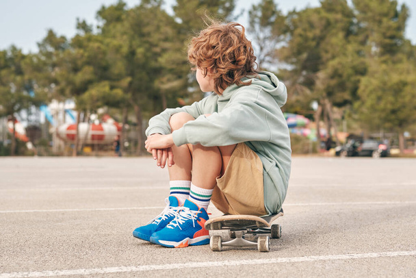 Vista laterale di anonimo teen boy in abito fresco seduto sullo skateboard e abbracciare le ginocchia in strada - Foto, immagini