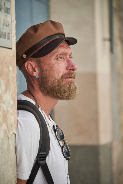 Πλευρική άποψη του γενειοφόρος αρσενικό τουρίστας σε καπέλο με σακίδιο στέκεται στο δρόμο και κοιτάζοντας μακριά με προσοχή - Φωτογραφία, εικόνα