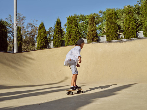 Boční pohled na chlapce bruslař v ležérní oblečení a s tenisky na koni skateboard na asfaltové silnici v slunečný den v parku se zelenými stromy pozadí - Fotografie, Obrázek
