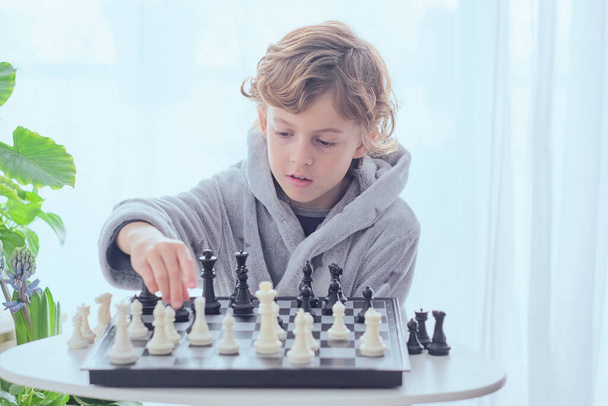 Geconcentreerde preteen jongen in grijs badstof badjas zitten aan tafel en schaken spel spelen in de lichte kamer - Foto, afbeelding