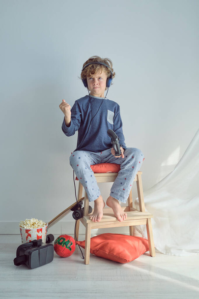 Full body of annoyed kid with blond hair in pajama and headphones playing video game with joystick sitting on wooden stool and clenching fist while losing game - Foto, Bild