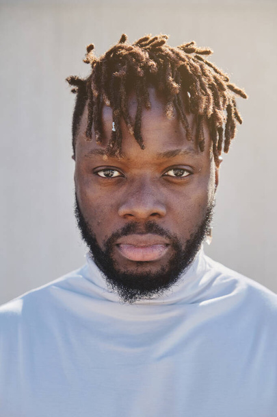 Unshaven African American guy with dreadlocks in white turtleneck looking at camera calmly on white backdrop - 写真・画像