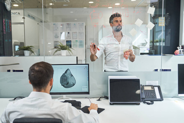 Обратный просмотр анонимного человека за компьютером и создание графической модели маски от пандемии коронавируса во время работы в офисе вместе с коллегой-мужчиной за стеклянной стеной - Фото, изображение