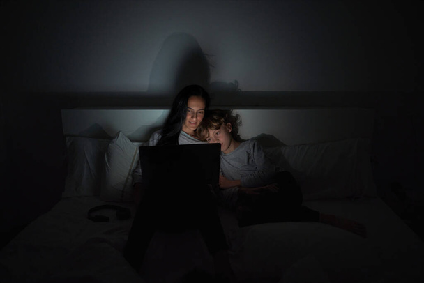 Kobieta z synem w casual nosić przeglądania laptopa podczas odrabiania pracy domowej razem na wygodne łóżko w ciemnej sypialni w domu - Zdjęcie, obraz