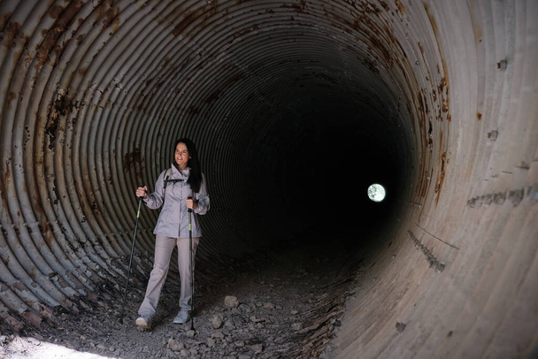Full body of active Hispanic female with walking poles standing in long dark tunnel with shabby rough walls during trip - Photo, Image