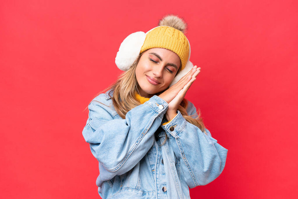 Young pretty Uruguayan woman wearing winter muffs isolated on red background background making sleep gesture in dorable expression - Photo, Image