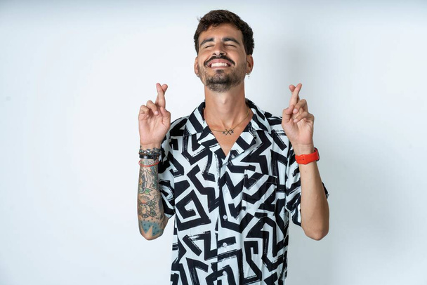 young caucasian man wearing printed shirt gesturing finger crossed smiling with hope and eyes closed. Luck and superstitious concept. - Photo, Image