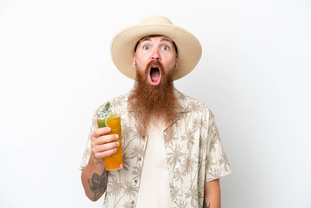 Uomo dai capelli rossi con lunga barba che beve un cocktail su una spiaggia isolata su sfondo bianco con espressione facciale a sorpresa - Foto, immagini