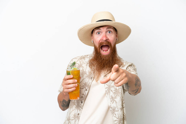 Homem ruivo com barba longa bebendo um coquetel em uma praia isolada no fundo branco surpreso e apontando frente - Foto, Imagem