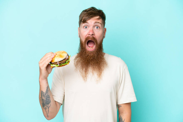 Pelirrojo con barba larga sosteniendo una hamburguesa aislada sobre fondo azul con expresión facial sorpresa y conmocionada - Foto, Imagen