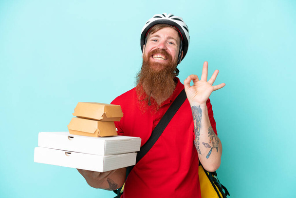 Liefermann hält Pizzen und Burger isoliert auf blauem Hintergrund und zeigt Ok-Zeichen mit Fingern - Foto, Bild