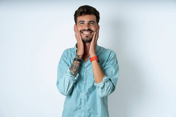 Happy handsome young man wearing turquoise shirt over white background touches both cheeks gently, has tender smile, shows white teeth, gazes positively straightly at camera, - Photo, Image