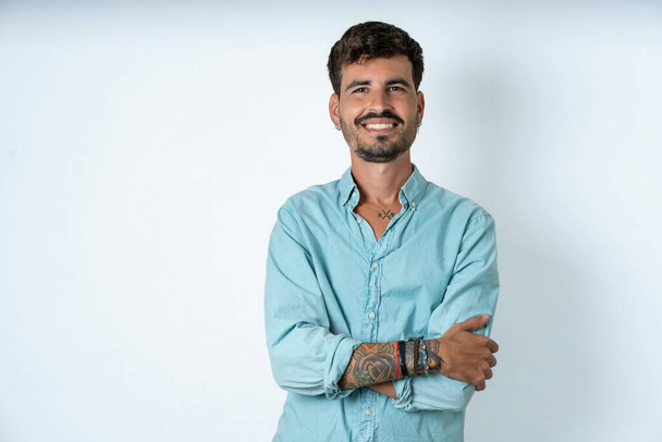 Portrét okouzlujícího pohledného mladého muže v tyrkysové košili na bílém pozadí stojí sebevědomě se zubatě usmívá s rukama složenýma - Fotografie, Obrázek