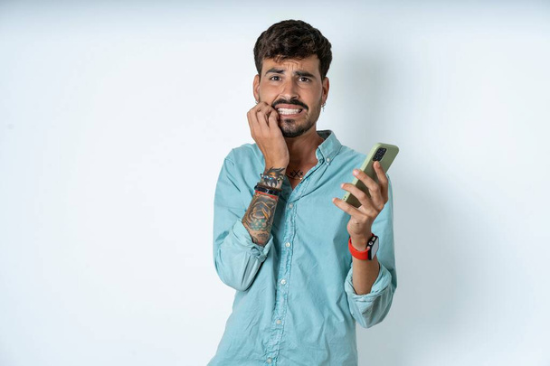 Peur drôle beau jeune homme portant chemise turquoise sur fond blanc tenant téléphone et ongles mordants - Photo, image