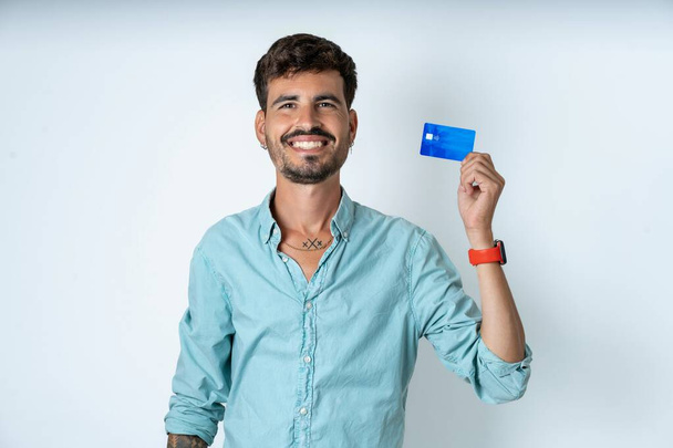 Foto veselý úsměv pozitivní pohledný mladý muž na sobě tyrkysové košile přes bílé pozadí doporučit kreditní kartu - Fotografie, Obrázek
