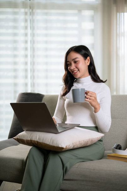 Красивая молодая азиатка в удобной одежде пользуется ноутбуком и пьет утренний кофе на диване в своей гостиной. Работа на дому, удаленная работа, фрилансер - Фото, изображение