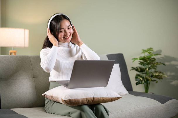 Kulaklık takan güzel bir Asyalı kadın, arkadaşıyla oturma odasındaki bir kanepede dizüstü bilgisayarından konuşuyor. Online toplantı, evden çalışmak, müzik dinlemek. - Fotoğraf, Görsel