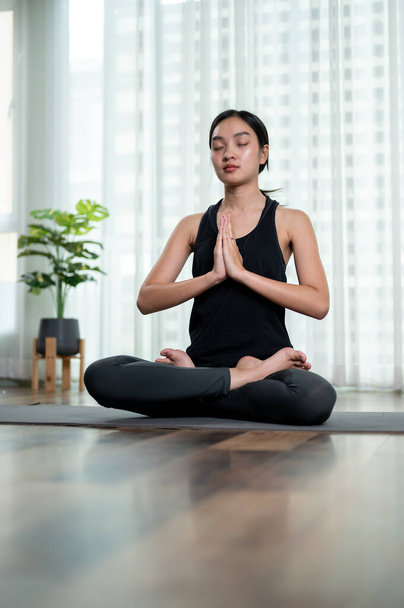 Une belle et calme asiatique en vêtements de sport médite sur un tapis de yoga dans son salon, pratiquant le yoga à la maison. entraînement de méditation, bien-être, mode de vie  - Photo, image