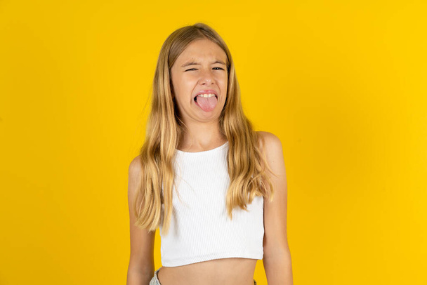 menina loira vestindo camiseta branca sobre fundo amarelo furando a língua feliz com expressão engraçada. Conceito de emoção. - Foto, Imagem