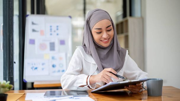 Una hermosa mujer de negocios musulmana asiática usando un hiyab está leyendo documentos, examinando el informe de negocios en su escritorio. - Foto, imagen