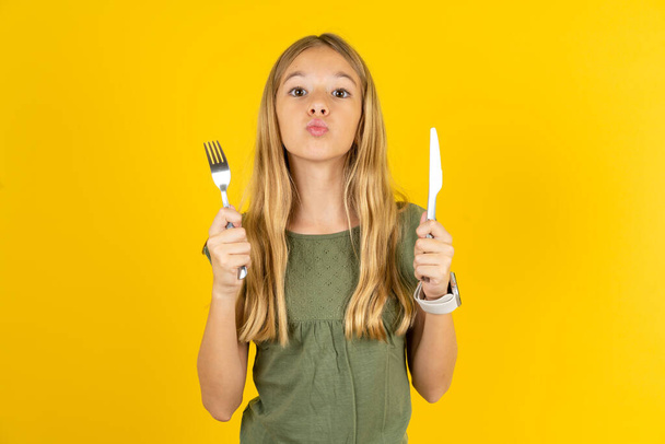 affamé blond petite fille portant chemisier kaki sur fond jaune tenant dans la main couteau fourchette veulent délicieuse pizza tarte - Photo, image
