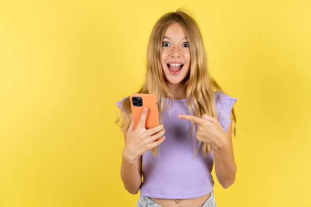 rubia chica vistiendo violeta camiseta sobre amarillo fondo celebración en las manos mostrando nueva celda, - Foto, imagen
